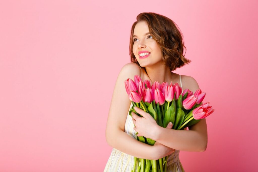cheerful-woman-with-bouquet-of-pink-tulips-looking-away-isolated-on-pink