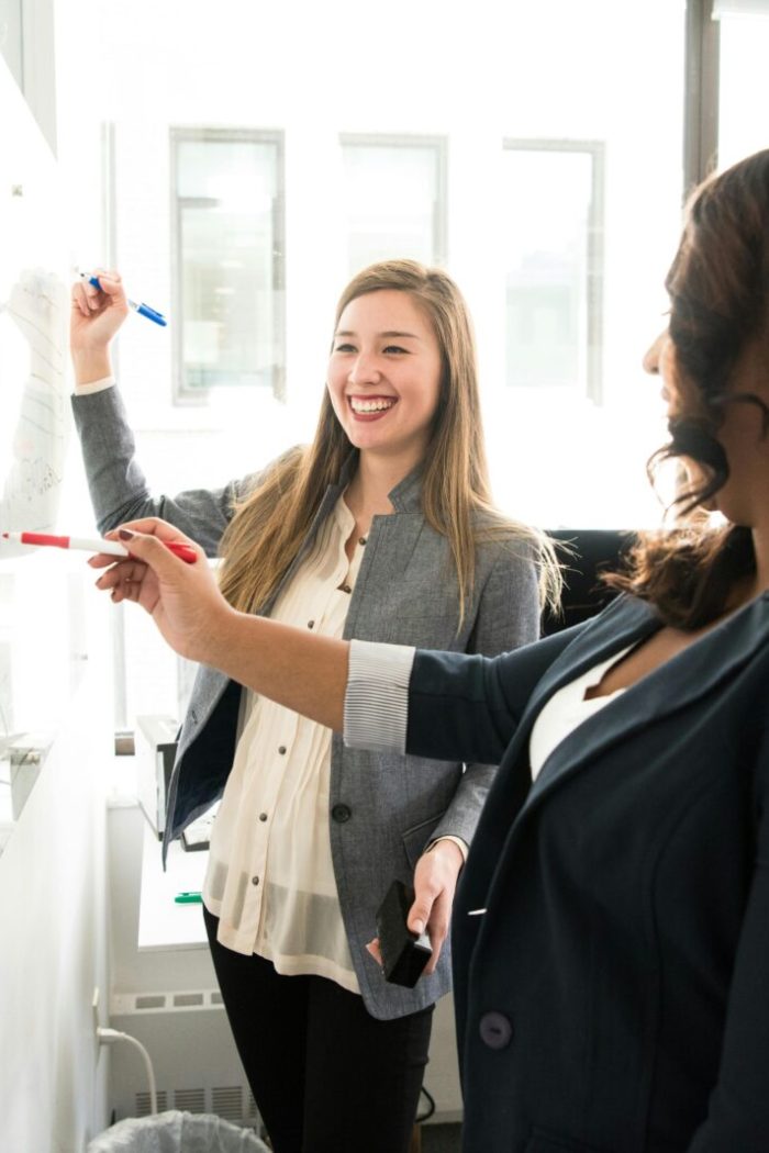 Two professional women discussing ideas on a whiteboard in a modern office setting.