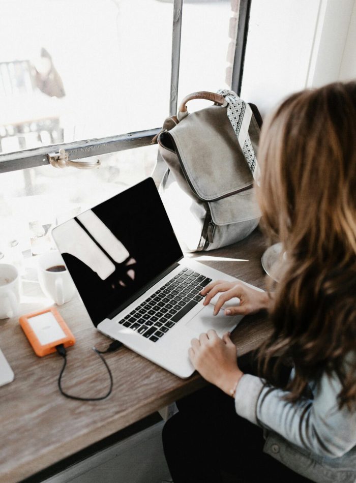 A young woman works remotely at a café, using her laptop and external hard drive.