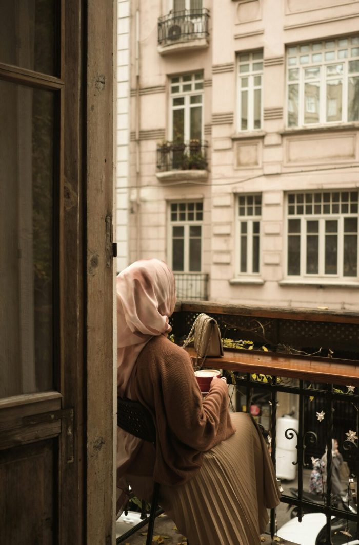 A woman in a hijab enjoys a peaceful moment on a balcony, observing the cityscape.