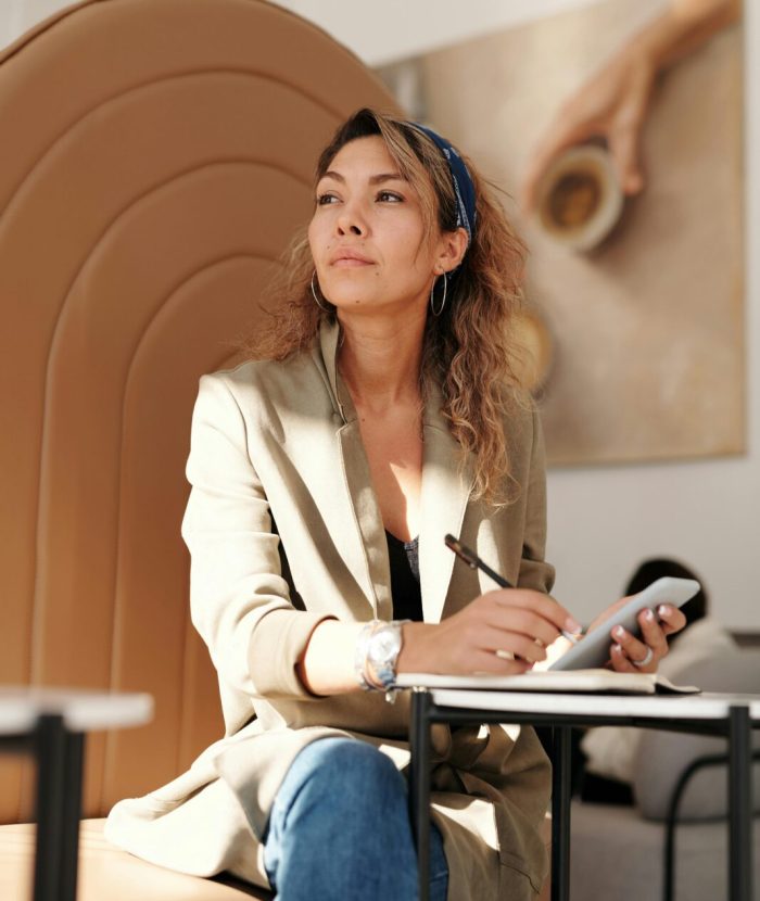 Confident woman in blazer taking notes with pen and smartphone in cafe.