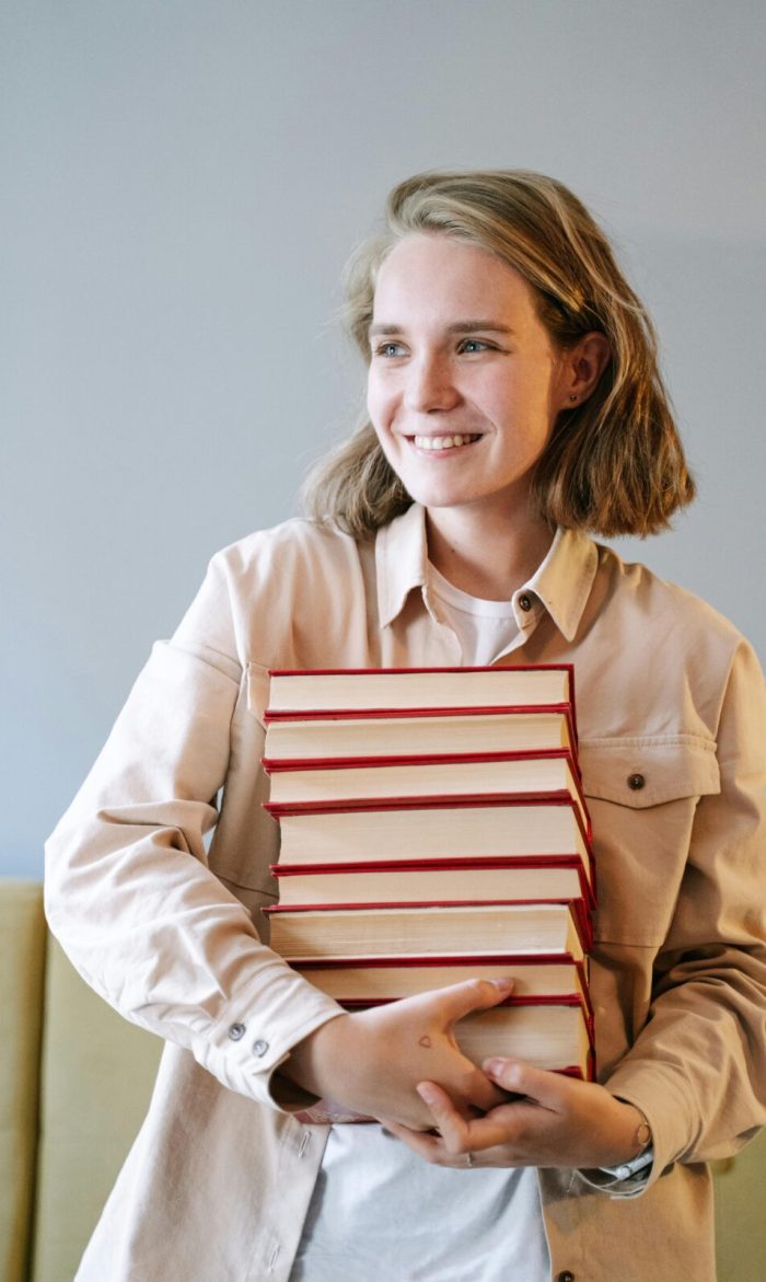 A young woman smiling while holding a stack of books indoors, embodying joy and education.