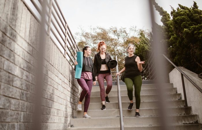 Three women walking down steps outdoors, carrying yoga mats, enjoying a fitness conversation.